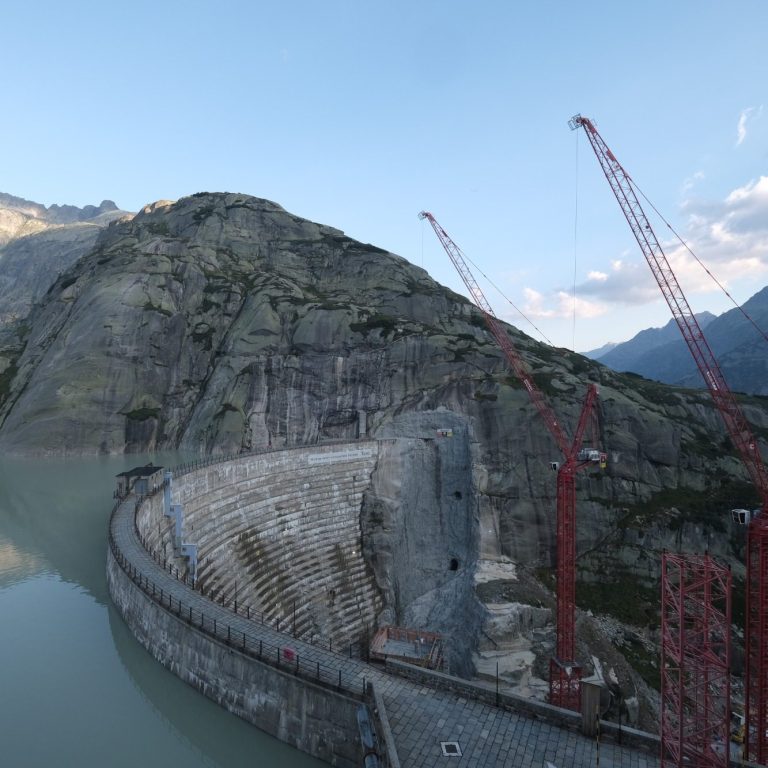 Staumauer in alpiner Umgebung bei klarem Wetter am frühen Morgen oberhalb der Krone. Baustelle mit Kränen.